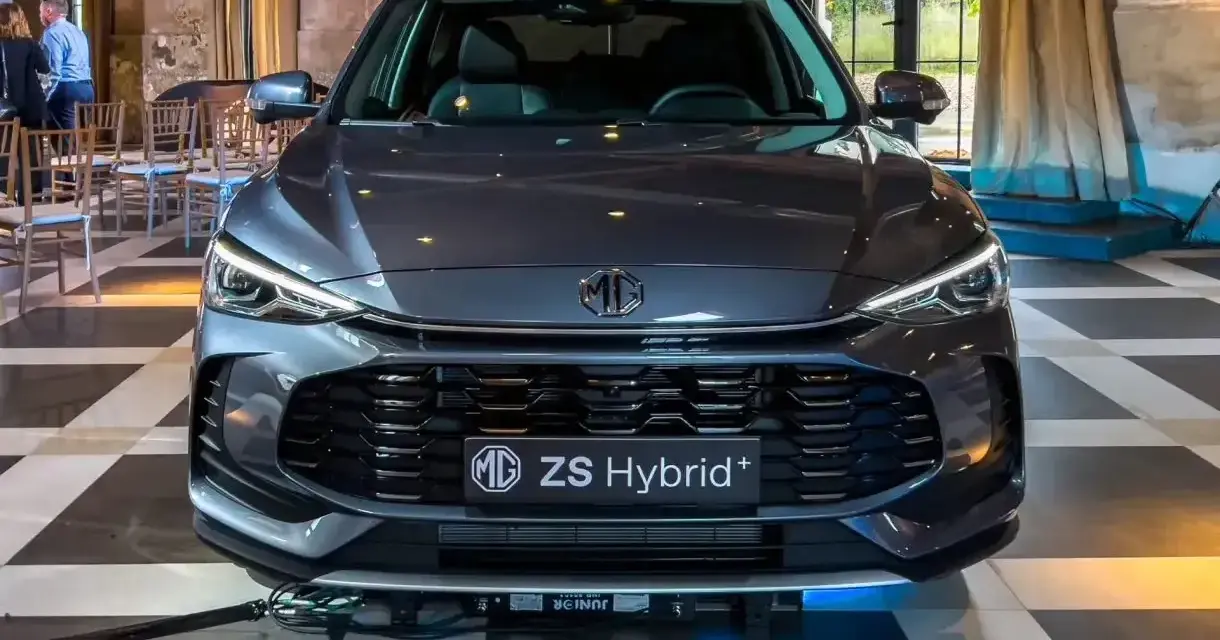 Szary MG ZS Hybrid+ prezentuje się elegancko na tle wnętrza z zasłonami i krzesłami.