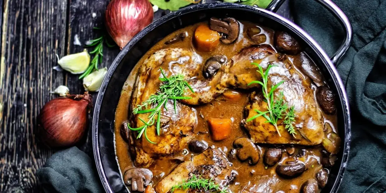 Herzhaftes Coq au Vin in einem Gusseisentopf, mit Pilzen, Karotten und Kräutern garniert. Daneben Zwiebeln und Knoblauch.