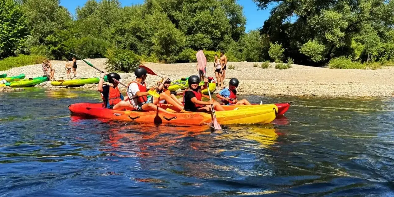 Des personnes profitent d'une activité canoe kayak sur une rivière ensoleillée, entourées de verdure et d'une plage de galets.