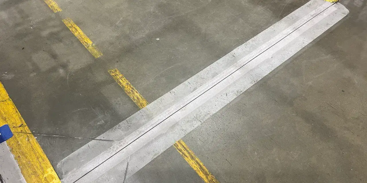 Dylatacja posadzki na betonowej podłodze, zaznaczona żółtymi liniami. Wygląda jak szew, który pozwala na ruchy konstrukcji.