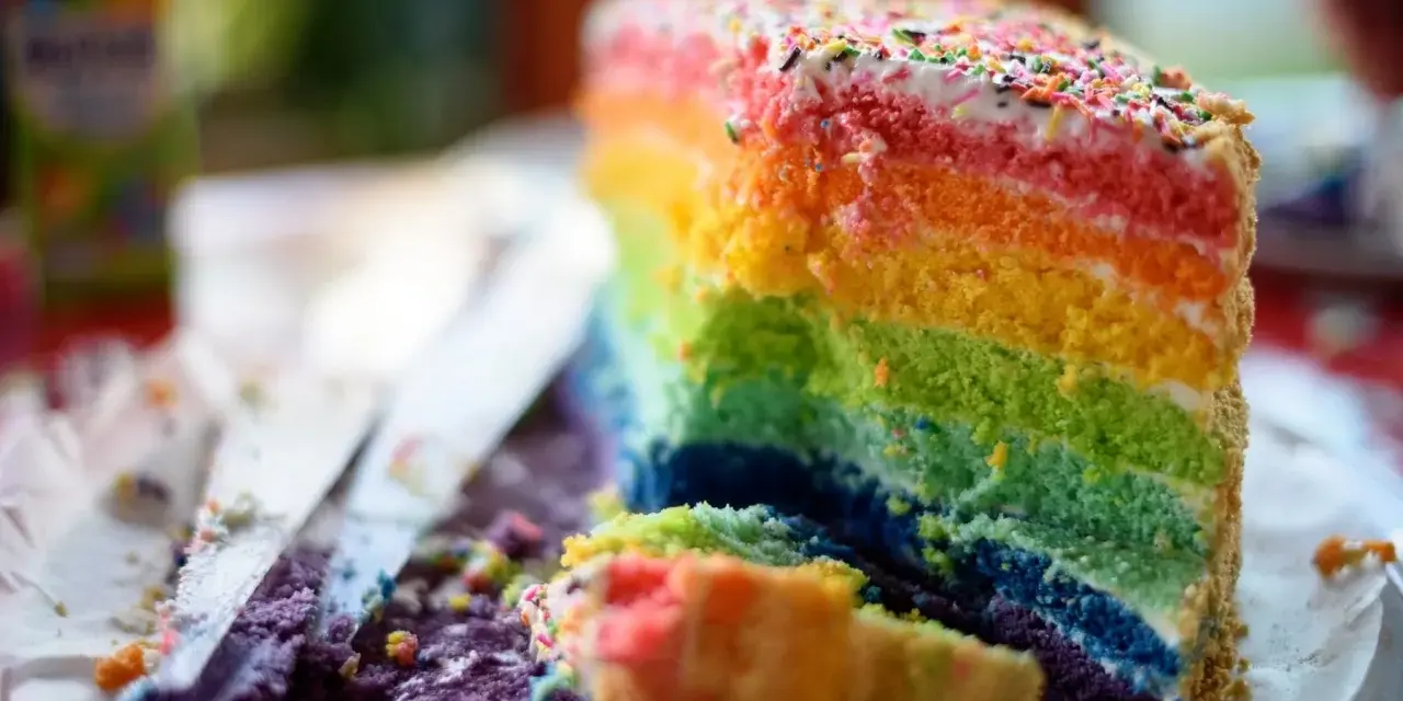Ein Stück bunter **Regenbogen Kuchen** mit bunten Streuseln, das auf einem Teller liegt. Daneben liegen Krümel und ein Messer.