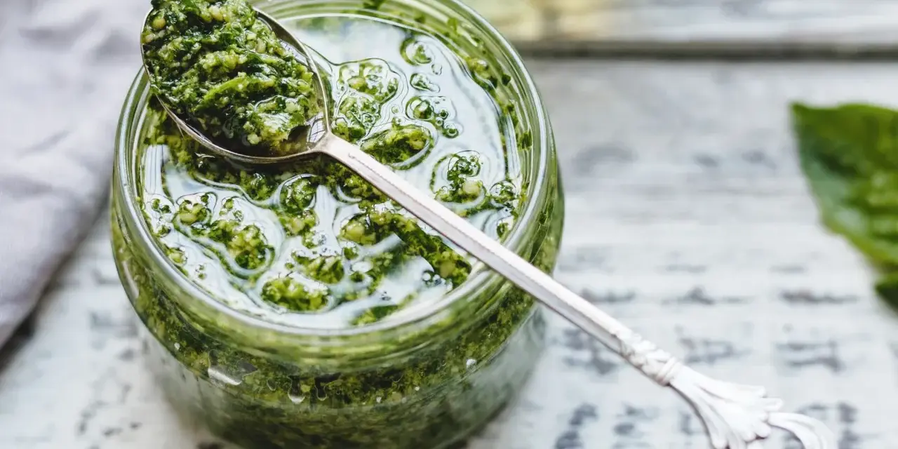Un vasetto di pesto genovese fatto in casa con frullatore, pronto per essere gustato. Un cucchiaino d'argento raccoglie un po' di questa delizia verde.