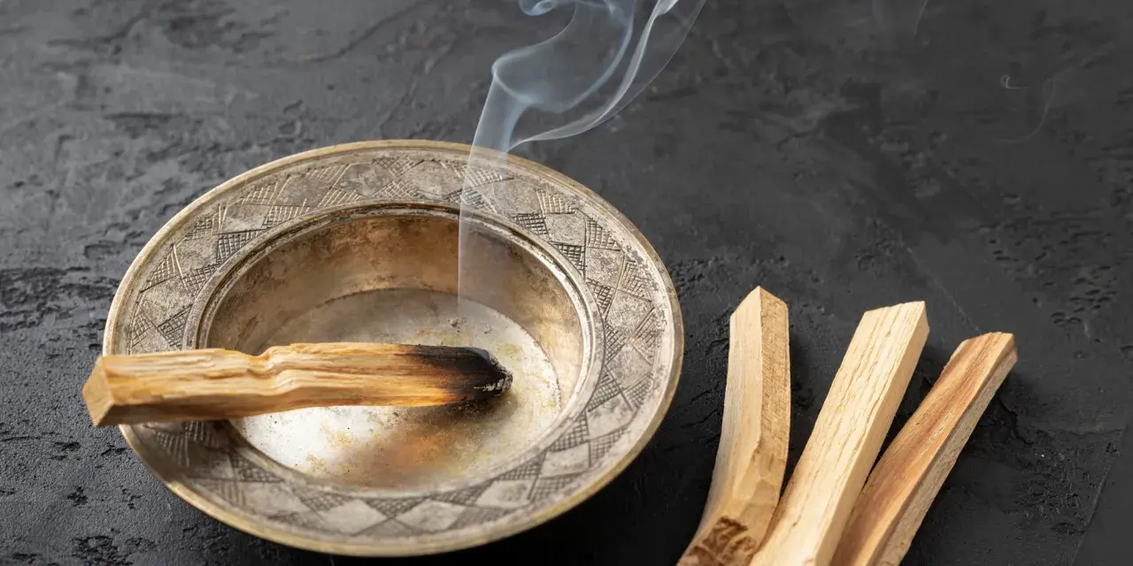 Un palo santo arde en un cuenco de metal, liberando humo blanco. A su lado, tres palos de palo santo sin quemar. Se puede quemar palo santo de noche para purificar el ambiente.