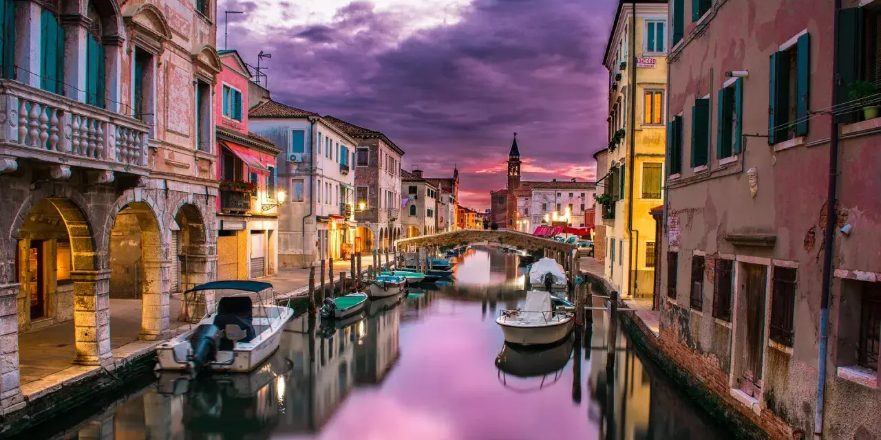Un canale di Venezia al tramonto, con barche ormeggiate e riflessi viola sull'acqua. Un'atmosfera magica che ricorda i piombi di Venezia.