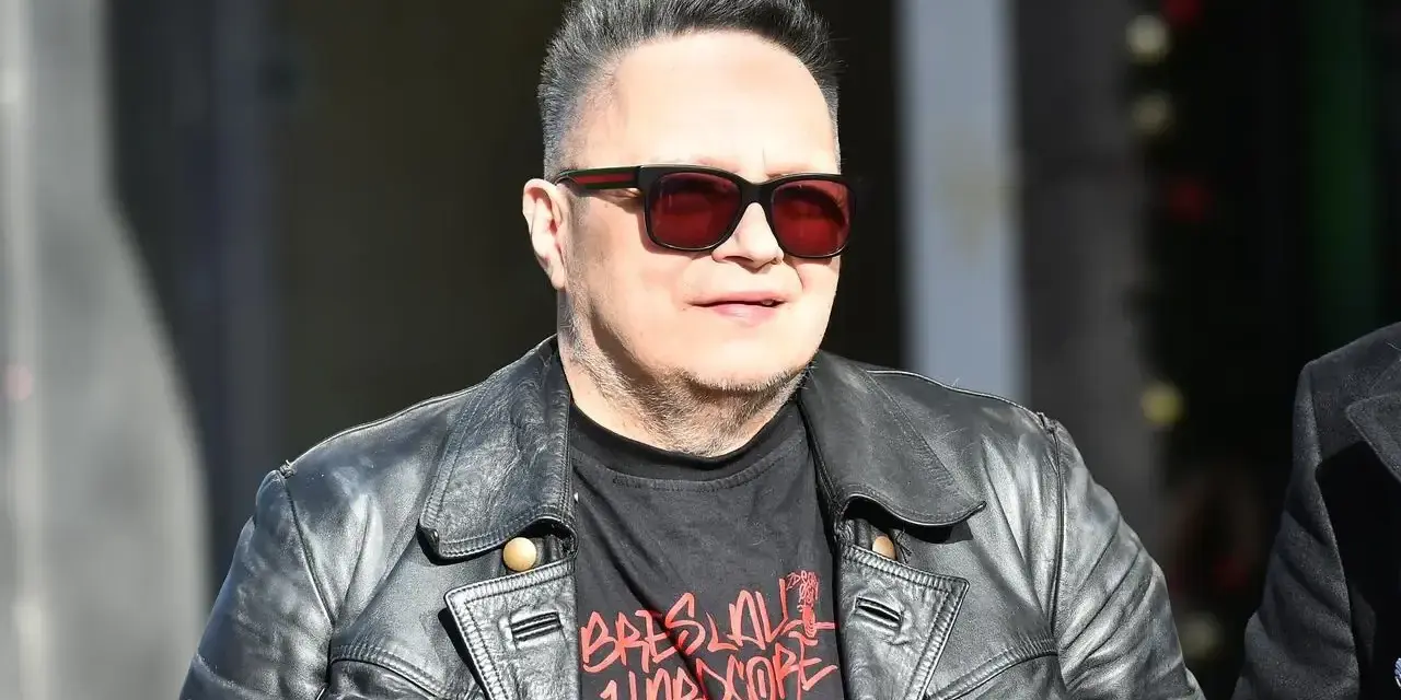 Charyzmatyczny wokalista w skórzanej kurtce i okularach przeciwsłonecznych, z charakterystyczną fryzurą.