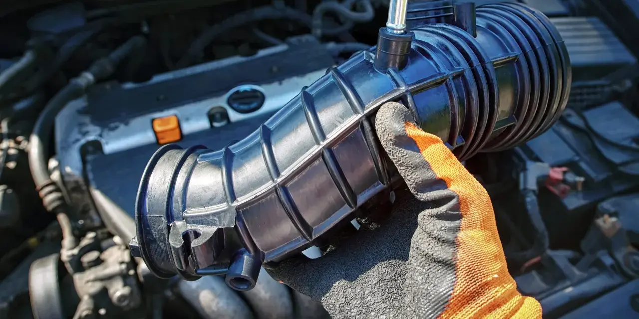 Gloved hand holds a black intake hose near a car engine. This part is often replaced during vacuum leak repair, affecting the vacuum leak repair cost.