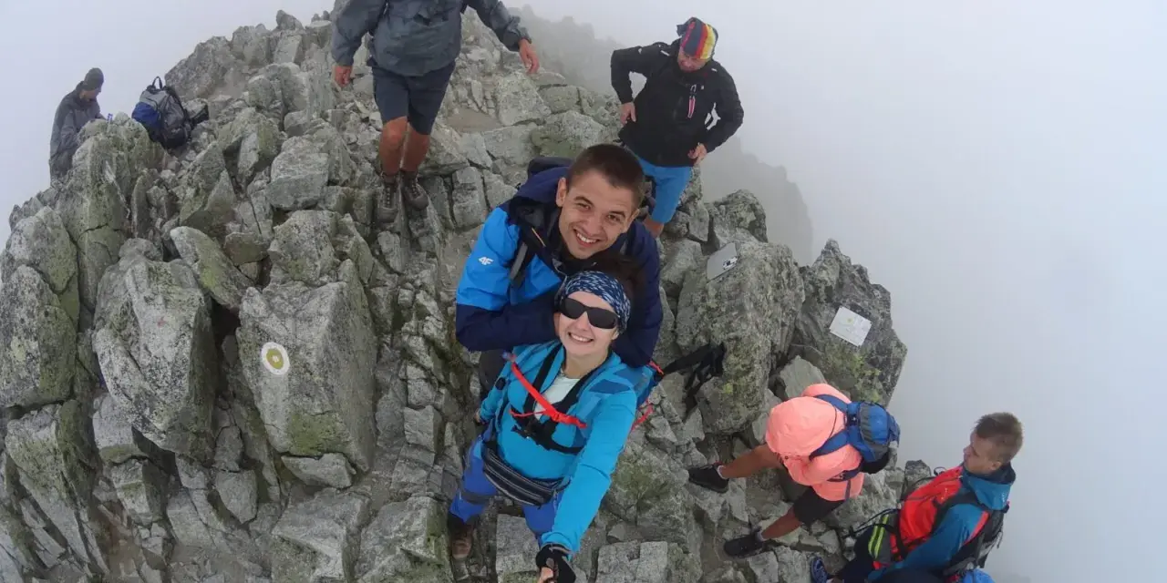 Grupa turystów na skalistym szczycie, otoczona mgłą. Jedna z kobiet, **mała wysoka**, robi sobie selfie z partnerem.