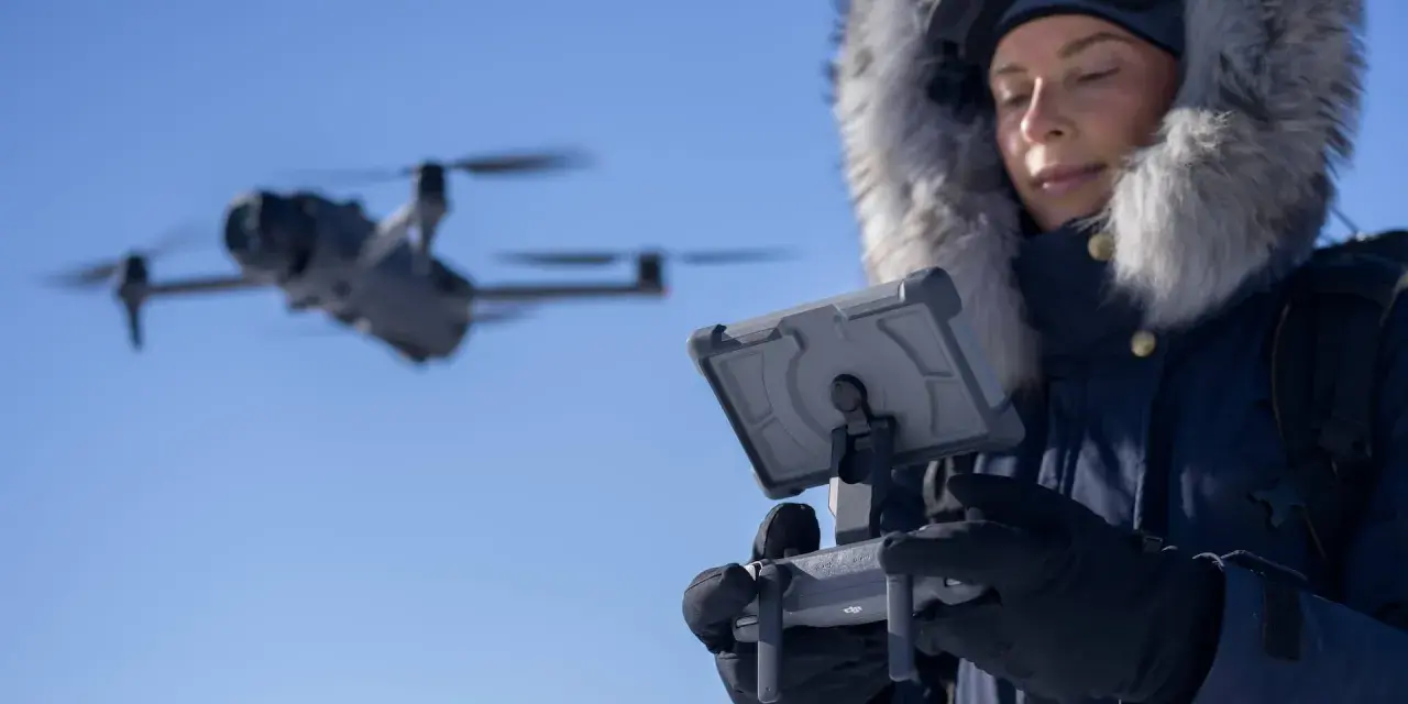 Woman in a fur-lined hood controls a flying drone in cold weather, looking at a tablet.