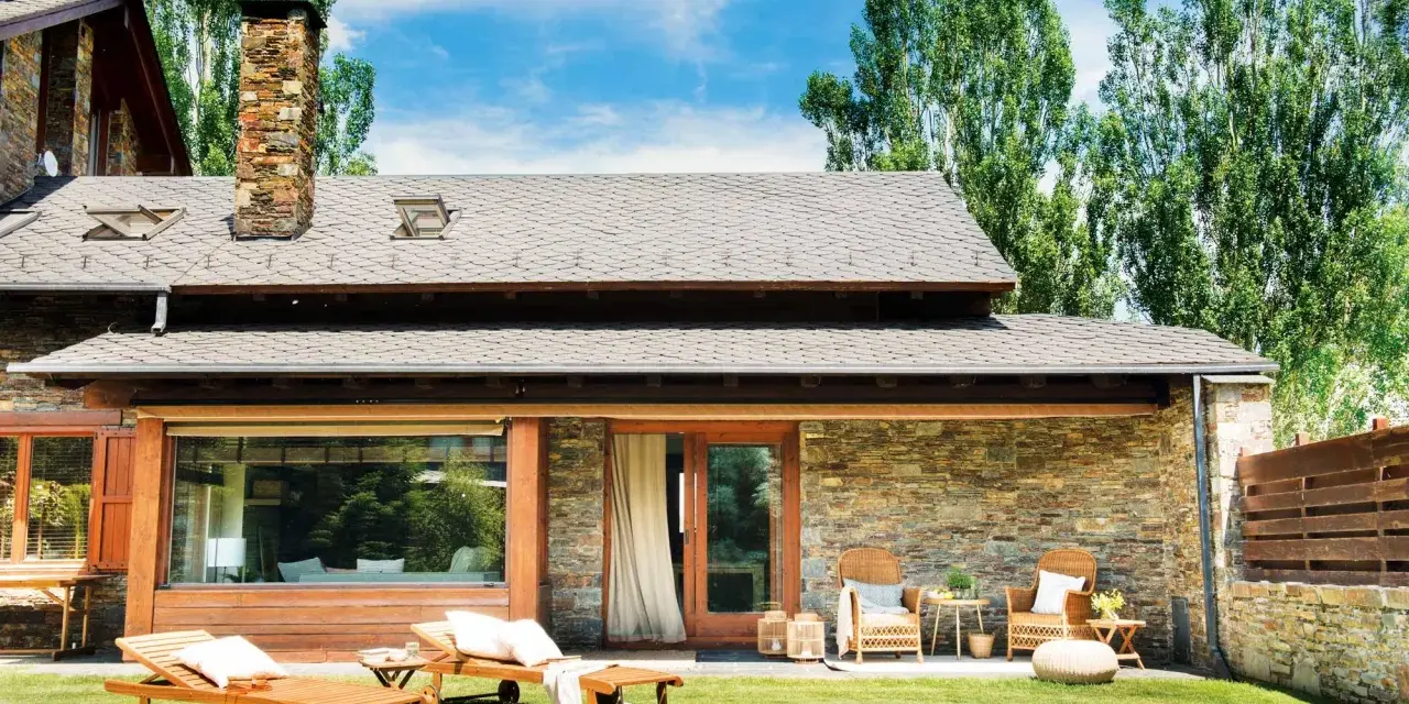 Fachadas de casas de pueblo modernas con piedra y madera, jardín con tumbonas y zona de estar exterior.