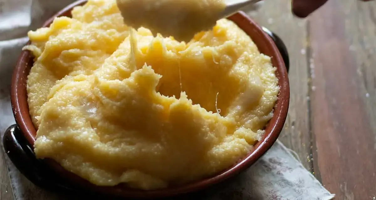 Un cucchiaio solleva una porzione filante di polenta concia valdostana, calda e cremosa, in una ciotola di terracotta.