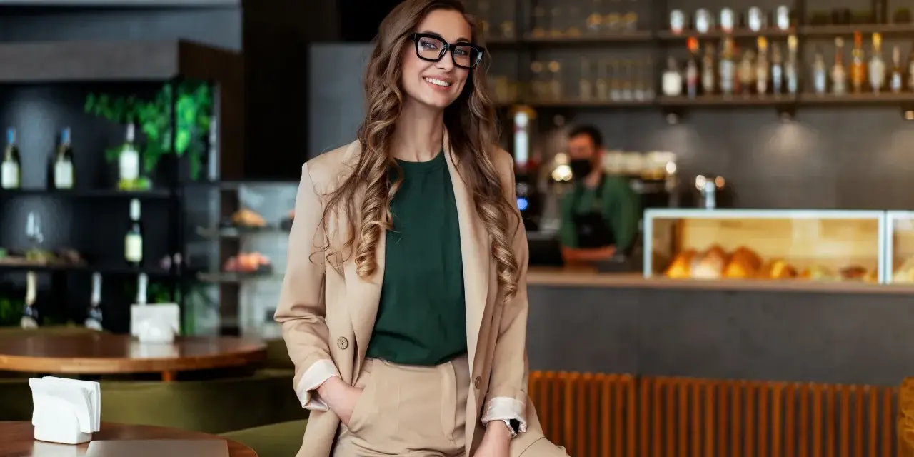 Mujer sonriente con gafas, vestida de traje beige, sentada en un restaurante moderno. Detrás, un barista trabaja. Esencial para la gestión de restaurantes.