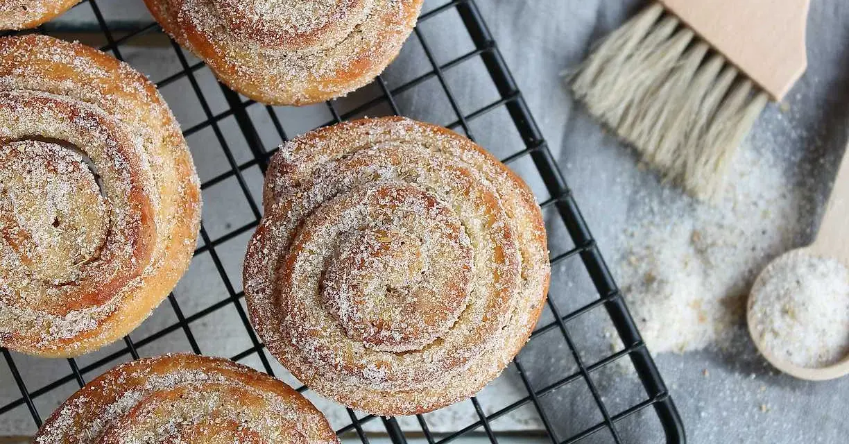 Nybakade karlsbaderbullar med socker och kanel på ett svart galler. En borste och en sked med socker ligger bredvid.