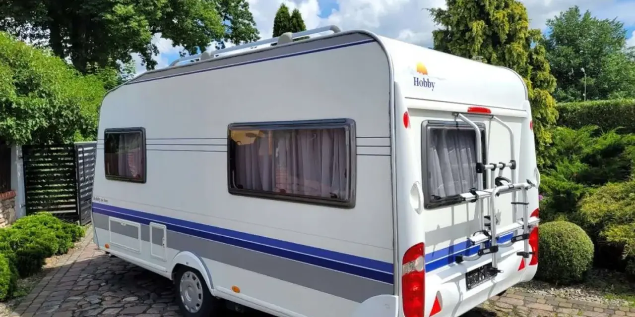 Biała przyczepa kempingowa Hobby 440 z niebieskimi pasami, zaparkowana na brukowanym podjeździe. Idealna do podróżniczego hobby.