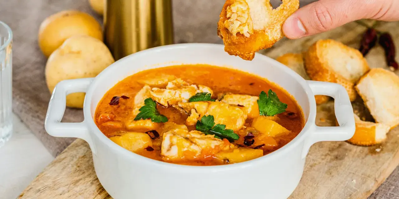 Una mano intinge un crostino nella zuppa di pesce surgelato, ricca di sapori e profumi.