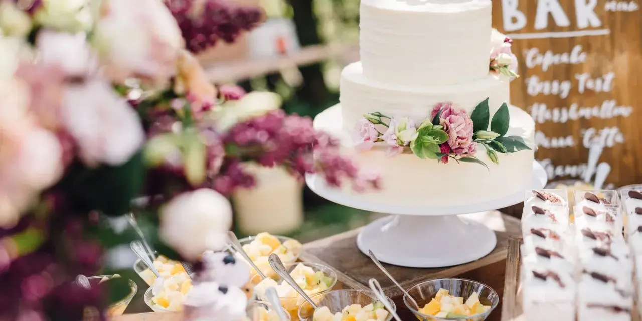 Elegancki tort weselny boho ozdobiony kwiatami, obok deserki i owoce.