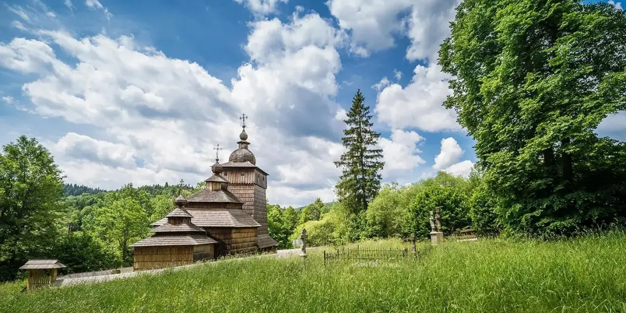Drewniana cerkiew na tle błękitnego nieba z chmurami, otoczona zielenią. Widok z łąki w Beskidzie Dolnym.