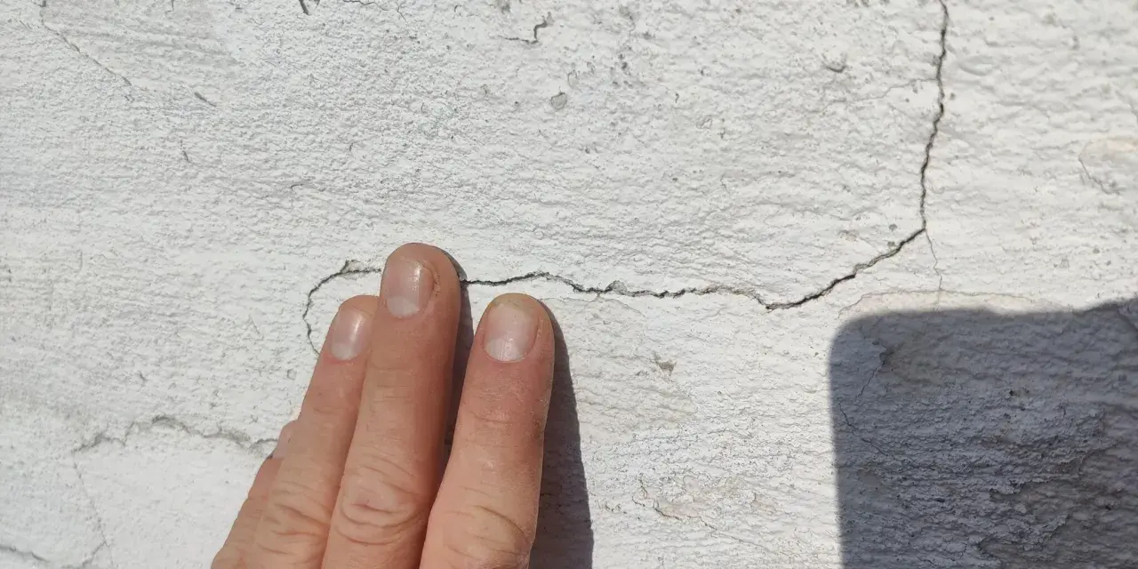 En hand pekar på sprickor i en vit, texturerad fasad.