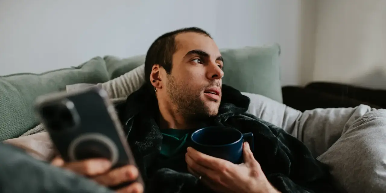 Mann liegt auf Sofa, hält Tasse und Handy. Mögliche Bewegungsmangel Symptome wie Müdigkeit und Lethargie werden angedeutet.