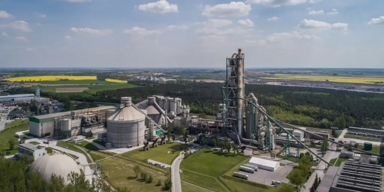 Zakłady Lafarge z lotu ptaka, z dominującą wieżą pieca cementowego i silosami, otoczone zielenią i polami.