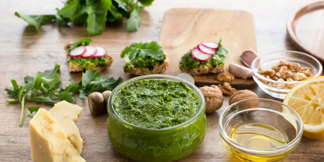 Zielone pesto w misce, obok składniki: ser, orzechy, cytryna, czosnek, oliwa.