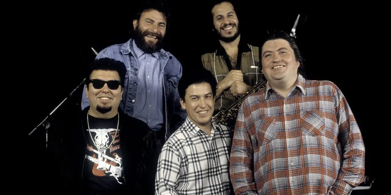 Los Lobos, native sons, pose together. One man wears sunglasses and a fringed jacket, another a denim jacket, and two sport plaid shirts. A saxophone is visible.