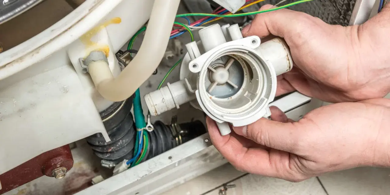 Hände halten eine Waschmaschine-Pumpe, die nicht abpumpt. Kabel und Schläuche sind sichtbar.