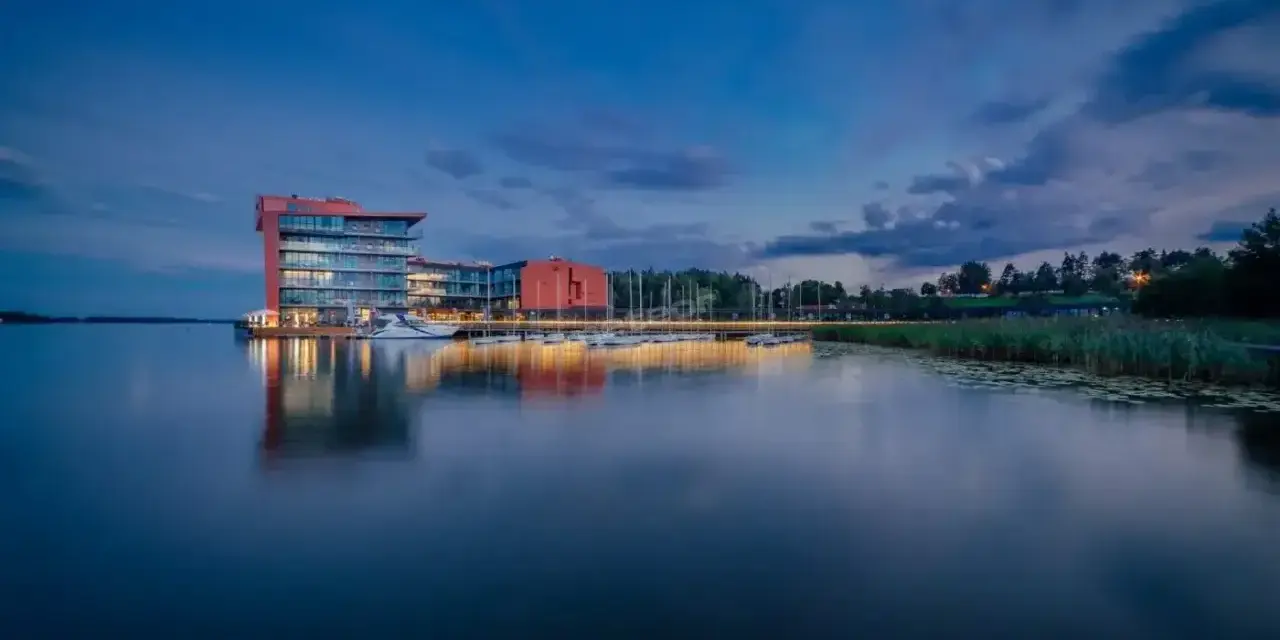 Nowoczesny hotel na ptasiej wyspie, otoczony spokojną wodą i zielenią, z łodziami przy pomoście.