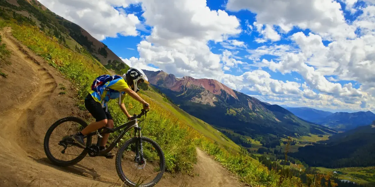 A mountain biker navigates a steep, winding trail, experiencing the best mountain biking in Colorado with stunning valley views and dramatic clouds.