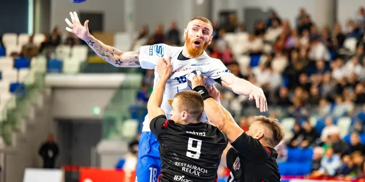 Jakub Wolski w akcji, walczy o piłkę w powietrzu z dwoma przeciwnikami.
