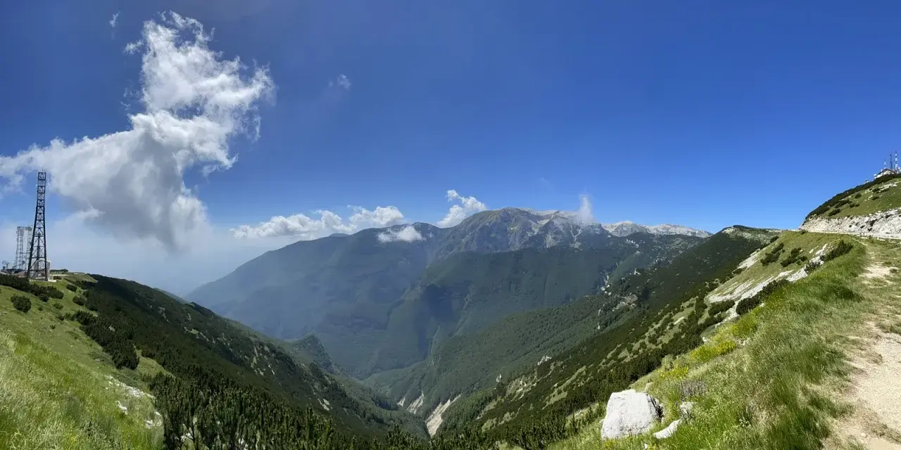 Panoramica mozzafiato sui monti dell'Abruzzo, con un sentiero che sale verso un **block house** e torri di trasmissione.