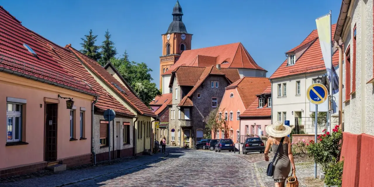 Frau mit Strohhut schlendert durch eine malerische Gasse mit Kopfsteinpflaster und roten Ziegeldächern. Ein besonderes Flair in Berlin.