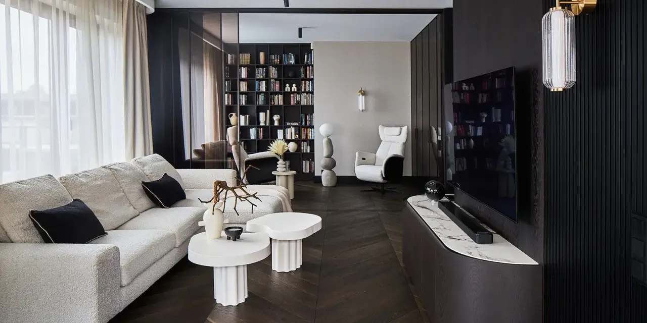 Nowoczesny salon z wygodną sofą, stolikami i regałem pełnym książek. Dominują ciemne, grafitowe akcenty, tworząc elegancką atmosferę.