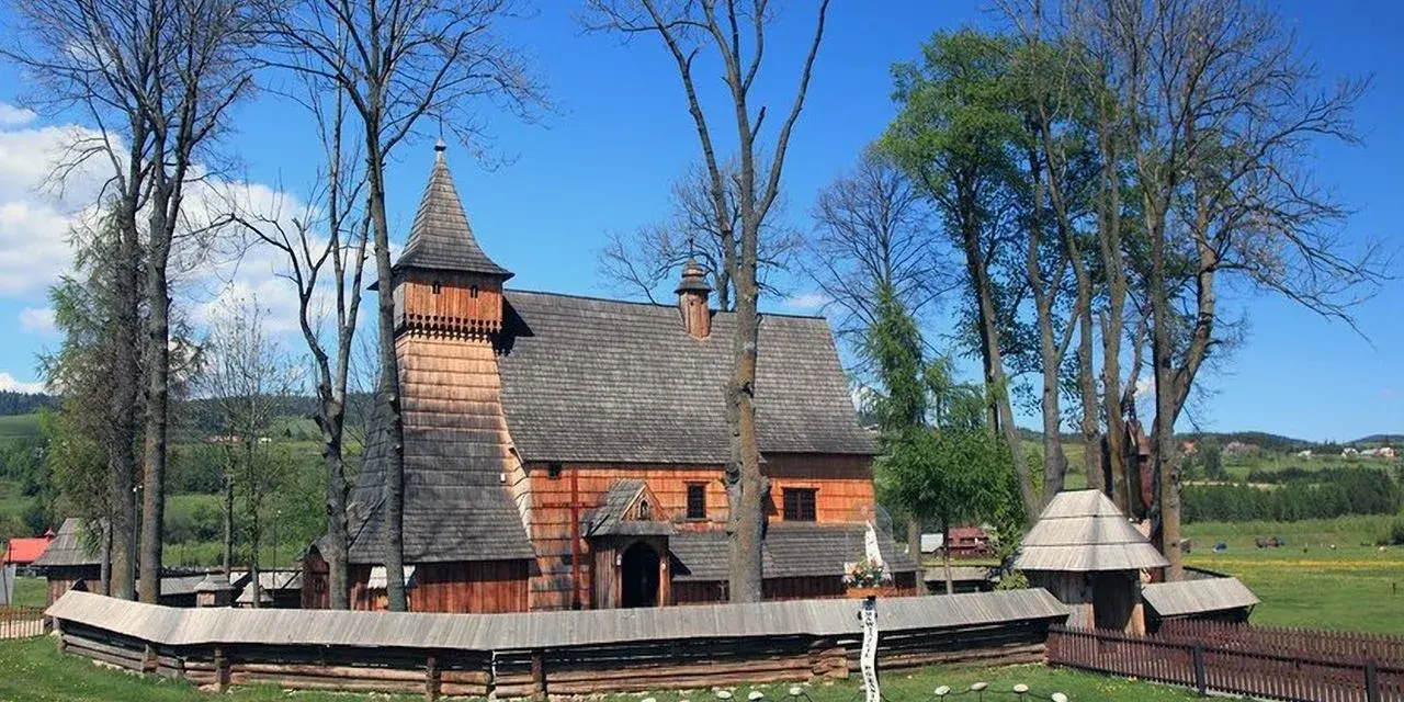 Drewniany kościół w Dębnie, otoczony kamiennym murem i drzewami, z krzyżem misyjnym na trawiastym dziedzińcu.