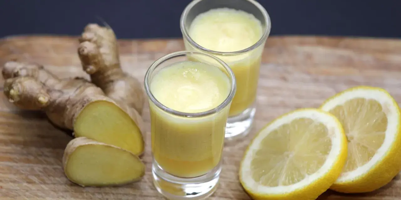 Zwei Gläser mit leuchtend gelbem Ingwer Shot, daneben frischer Ingwer und Zitronenscheiben auf Holzbrett.