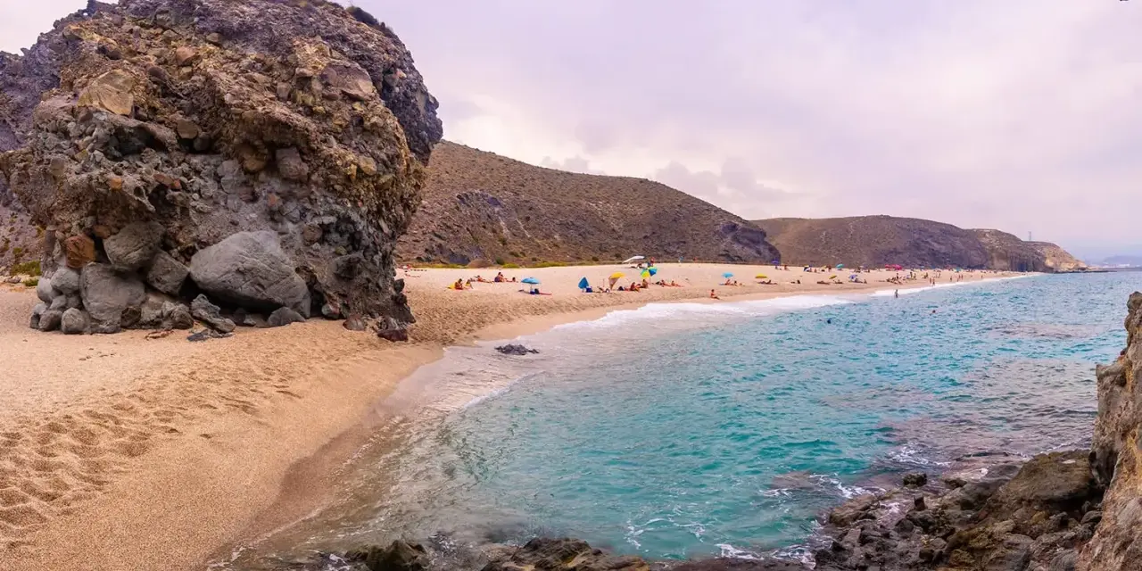 Plaża w Cabo de Gata z turkusową wodą, piaszczystym brzegiem i skalistymi formacjami. Ludzie odpoczywają pod parasolami.