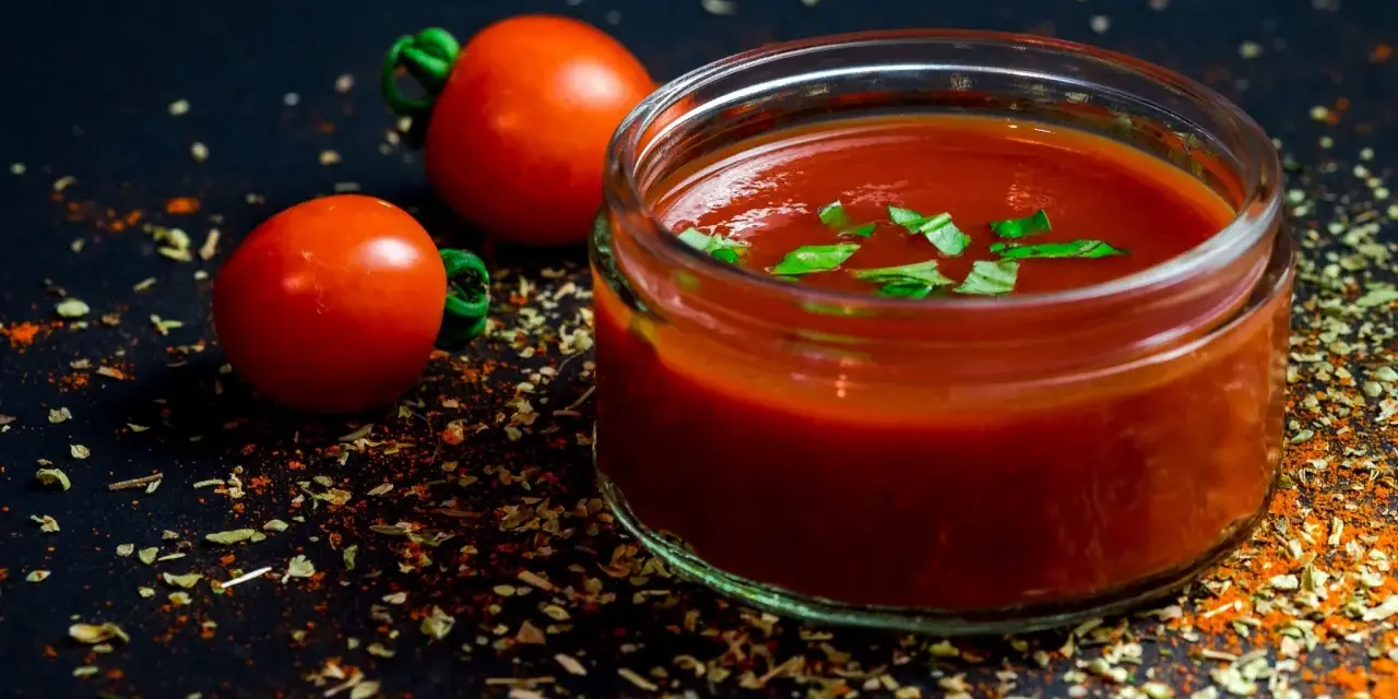 Un vasetto di ketchup fatto in casa, guarnito con basilico fresco, accanto a pomodori maturi e spezie su sfondo scuro.