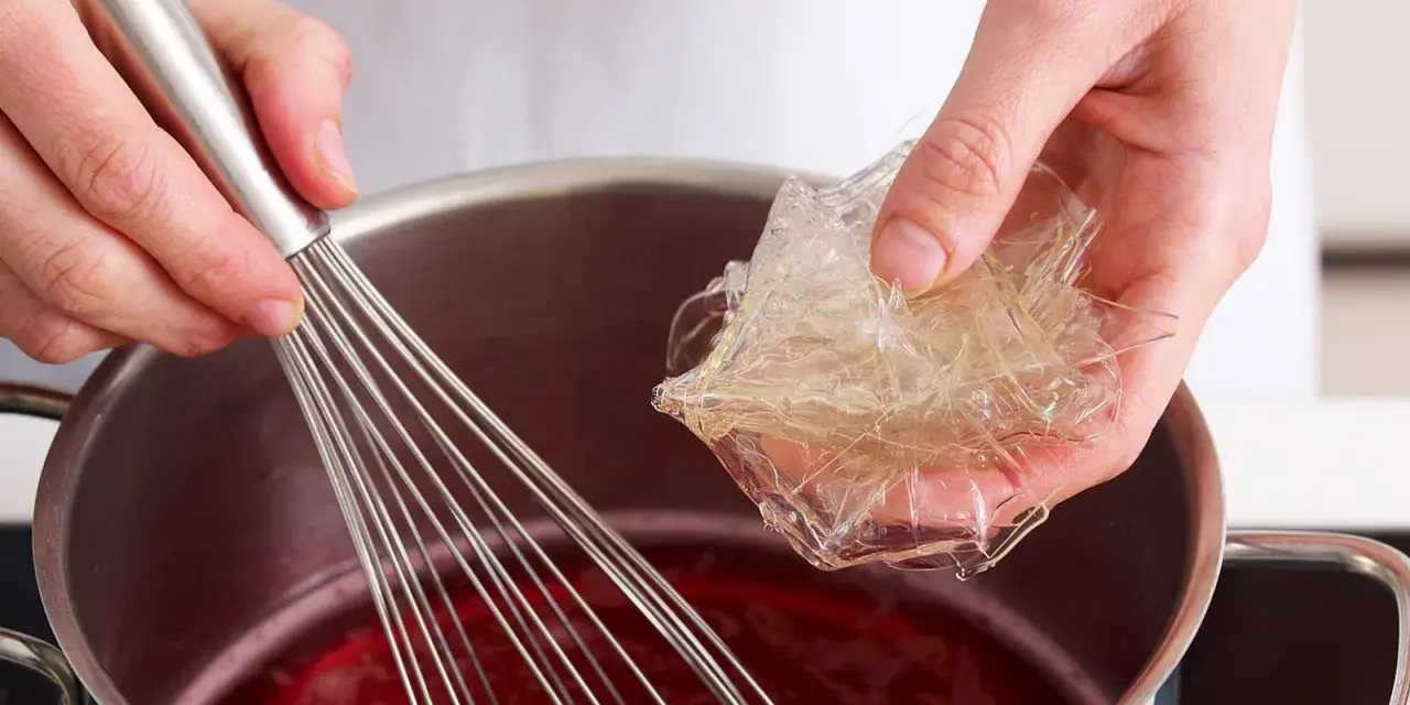 Hände bereiten Gelatine-Blätter für die Zubereitung vor, indem sie sie in eine rote Flüssigkeit in einem Topf geben, während ein Schneebesen bereitsteht.