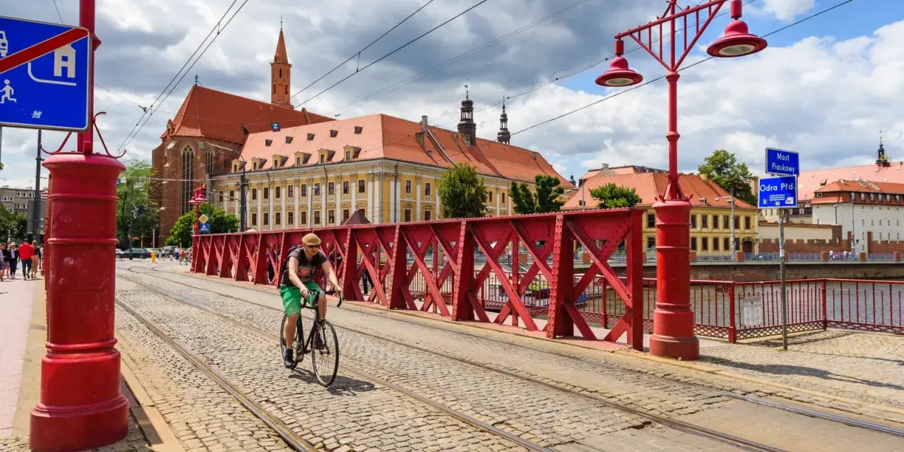 Mężczyzna na rowerze jedzie po brukowanej drodze rowerowej z torami tramwajowymi, w tle zabytkowe budynki i czerwony most.