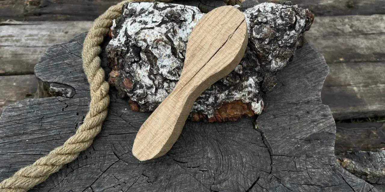 Cucchiaio grezzo in legno d'acero, appoggiato su un tronco scuro con una corda rustica.