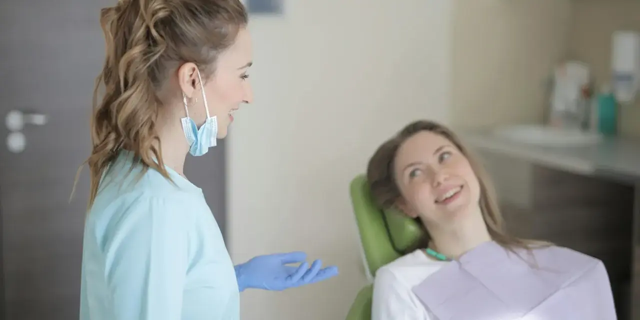 Uśmiechnięta pacjentka na fotelu stomatologicznym słucha dentystki. To obrazek z gabinetu, gdzie odbywają się studia stomatologiczne.