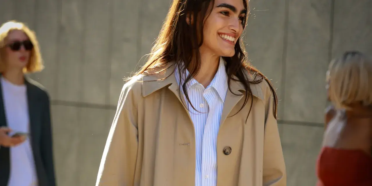 Donna sorridente con capelli lunghi e castani indossa un trench beige aperto su una camicia a righe azzurre. Perfetto per ogni occasione, anche se posso mettere il trench di sera.