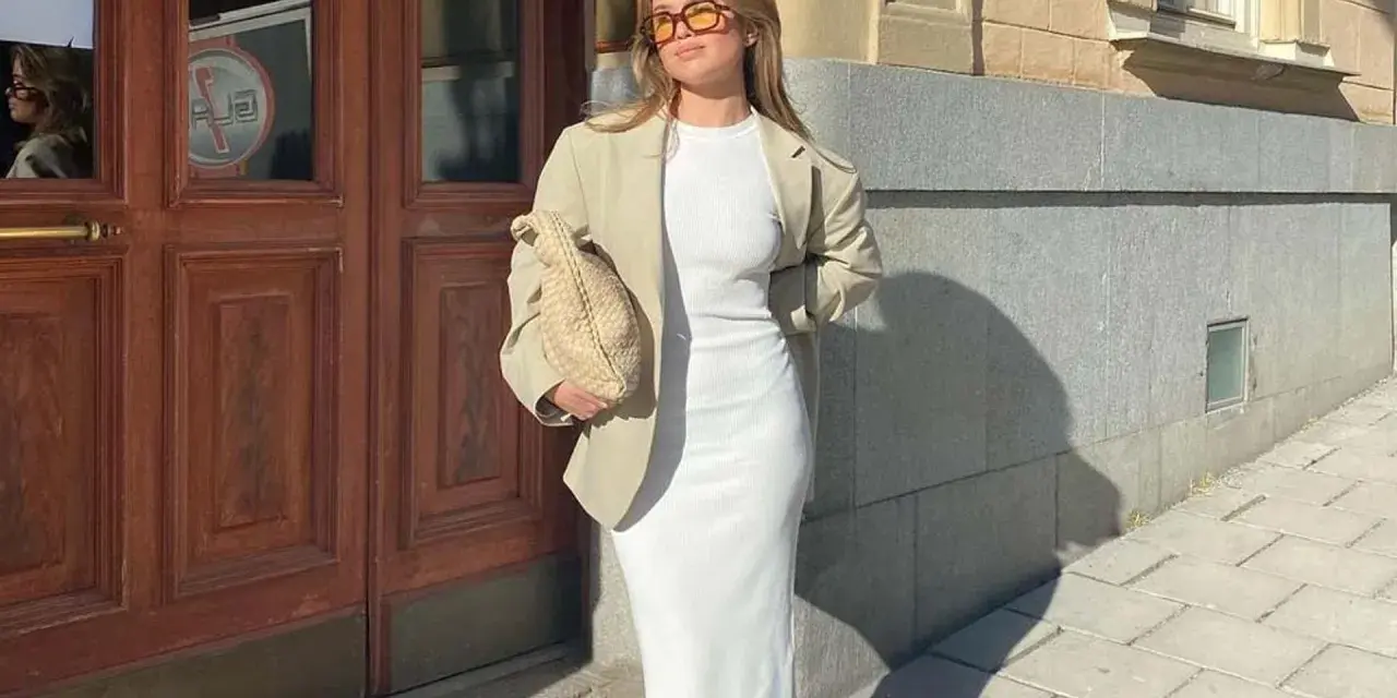 Mujer con blazer y vestido largo blanco, bolso tejido y gafas de sol, posando frente a una puerta de madera.
