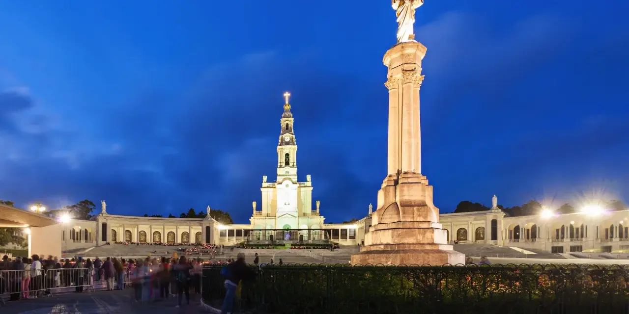 Pilgrims gather at the Sanctuary of Fátima, Portugal, at dusk, with the illuminated basilica and statue of Christ the King standing majestically.