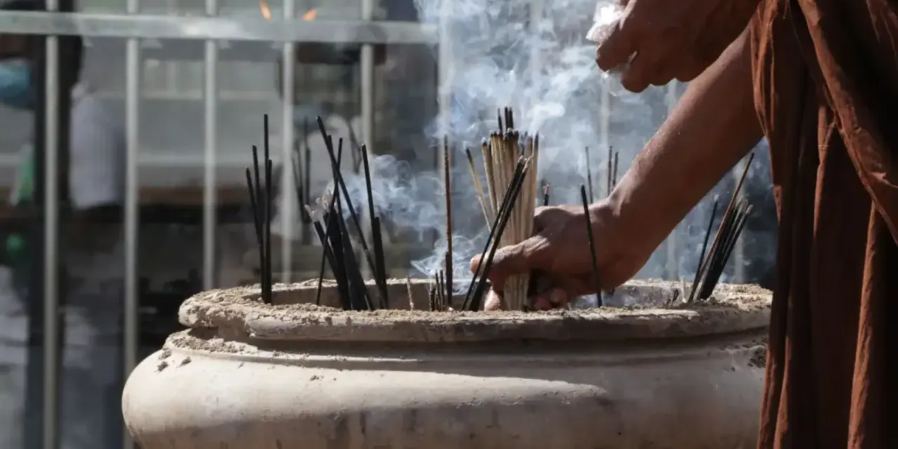 Manos monje en túnica marrón colocando varillas de incienso en un cuenco de piedra, creando humo para limpiar el ambiente.