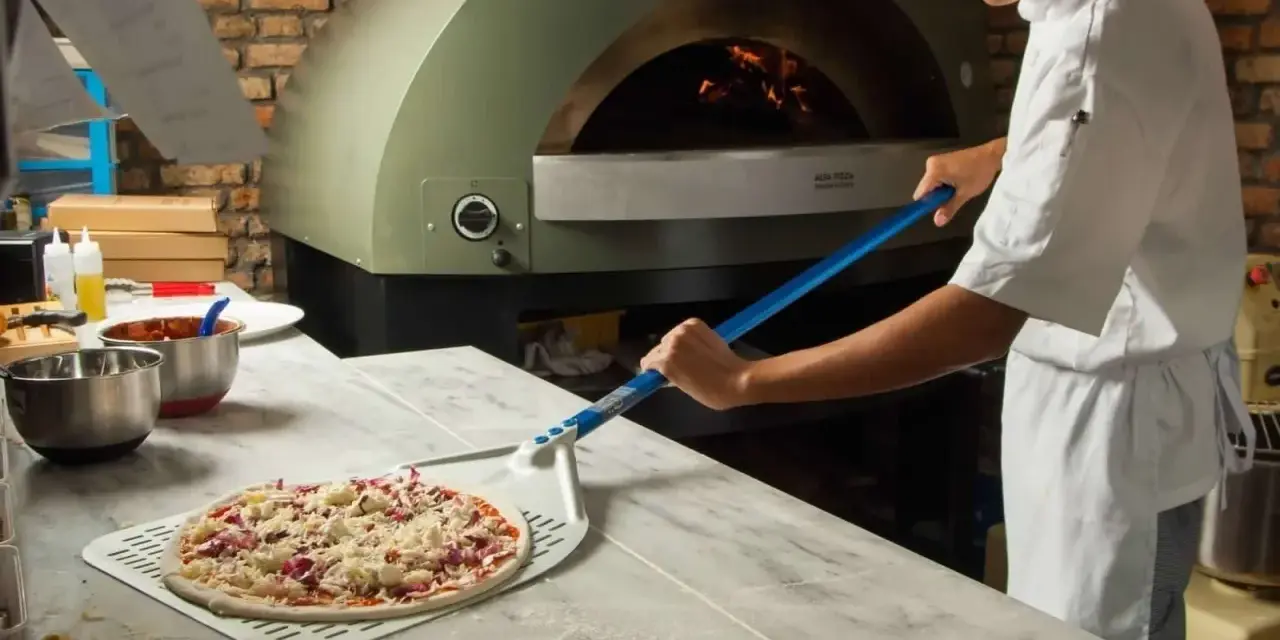 Pizza cruda en pala, lista para horno de leña con chef al fondo.