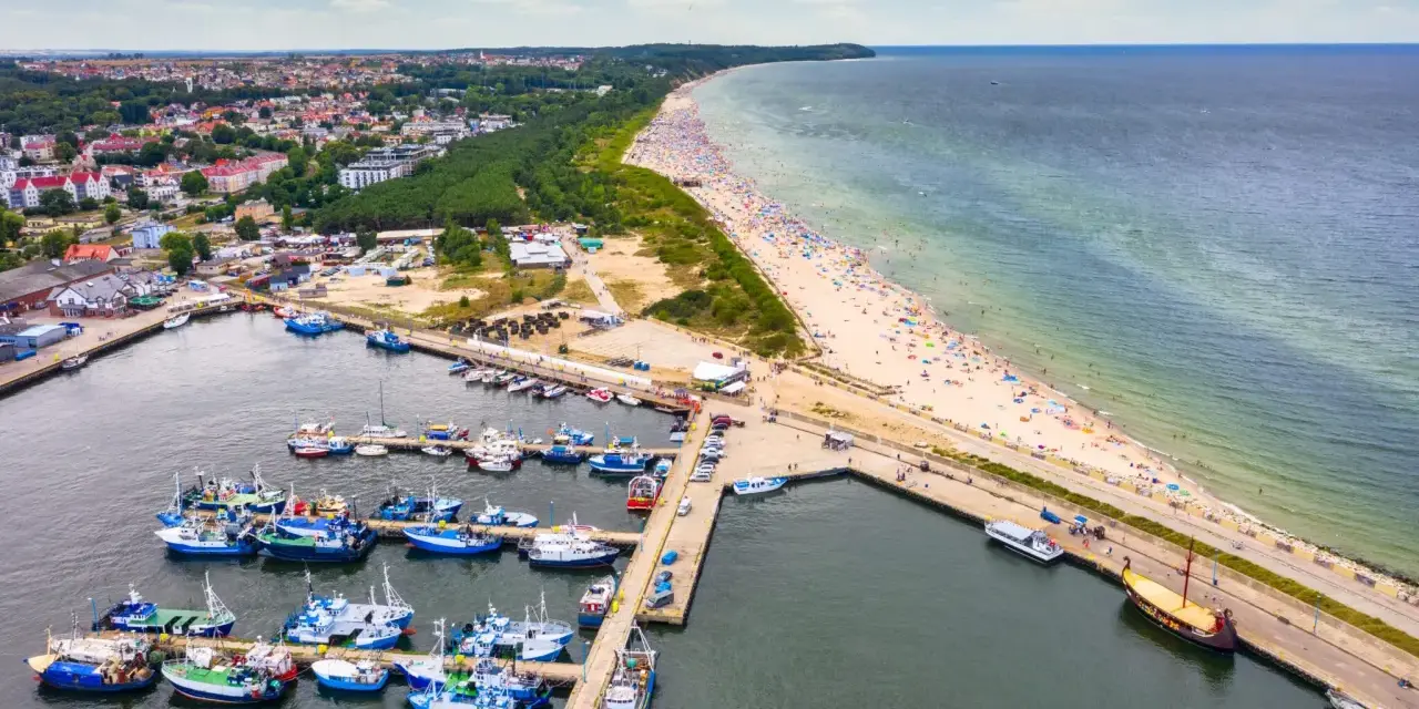 Port rybacki we Władysławowie, województwo pomorskie, tętni życiem. W tle rozciąga się plaża pełna turystów i morze.