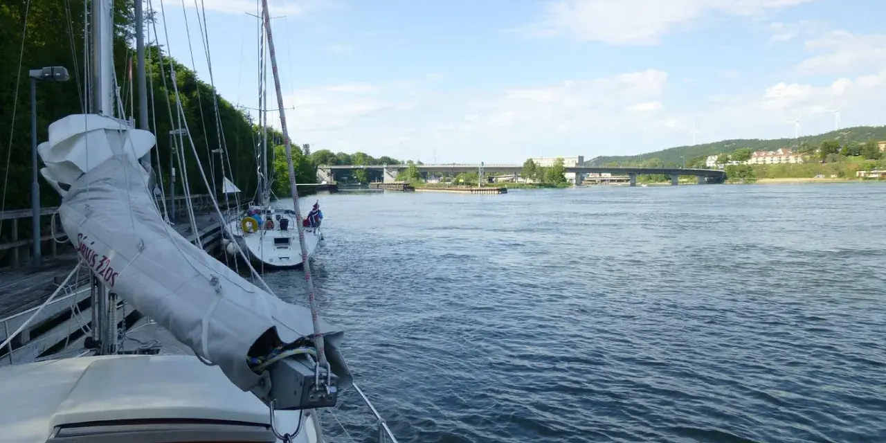Segelbåtar vid kaj i Lilla Edet, en bro syns i bakgrunden över en bred flod.