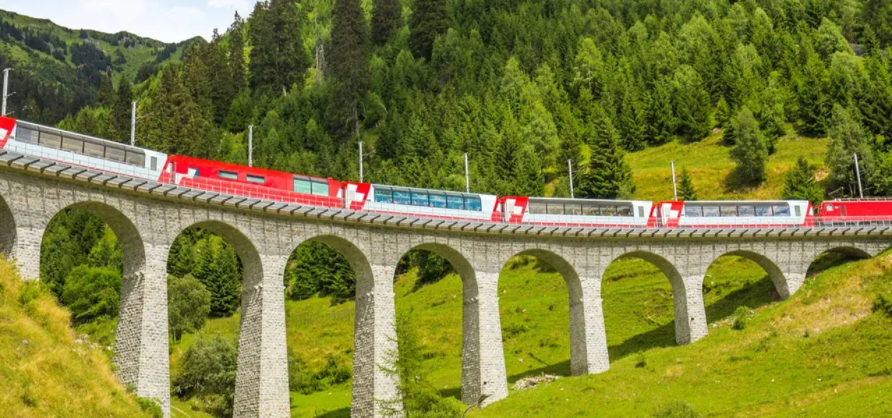 A red and white train travels across a stone viaduct, winding through a lush green valley with a dense forest on the mountainside. This scenic route is part of the Amsterdam to Basel train journey.