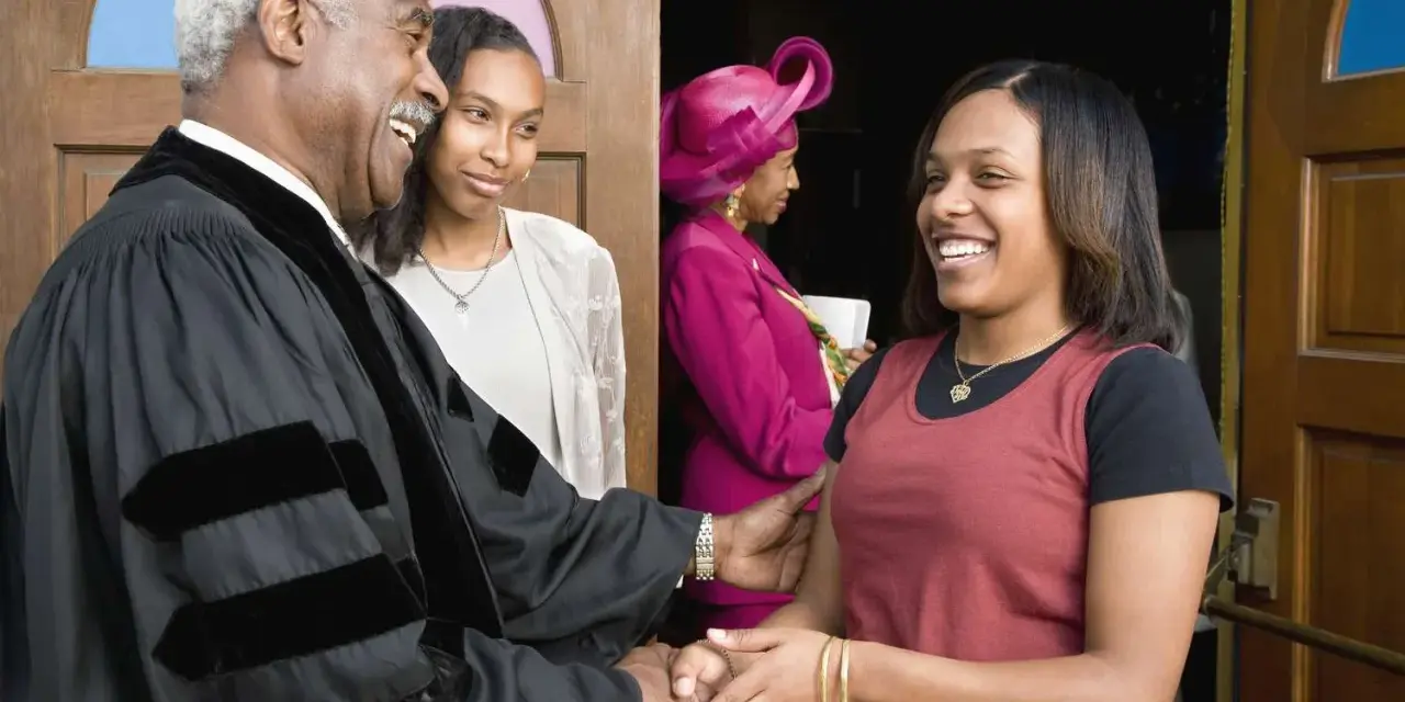 A deacon in the church, wearing robes, greets a young woman with a handshake.