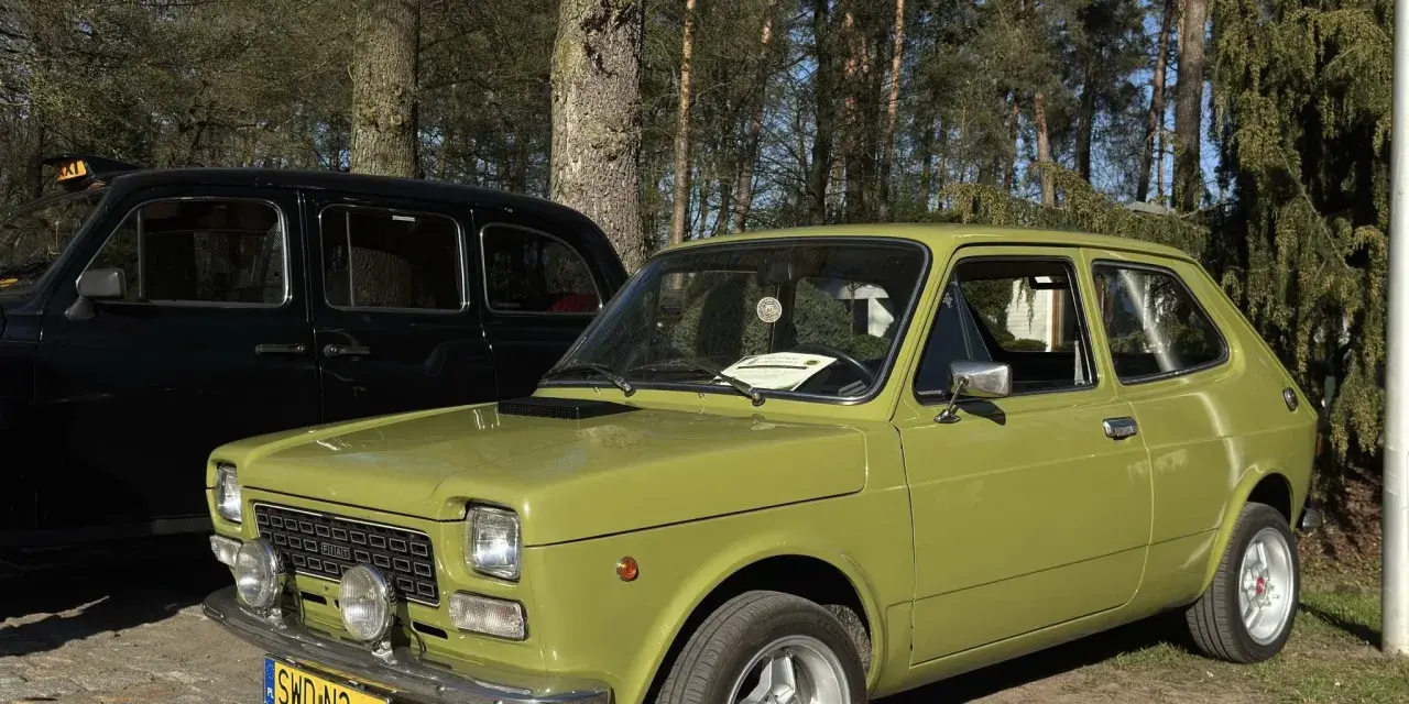 Zielony fiat 127 z lat 70. stoi obok czarnej taksówki na tle drzew.